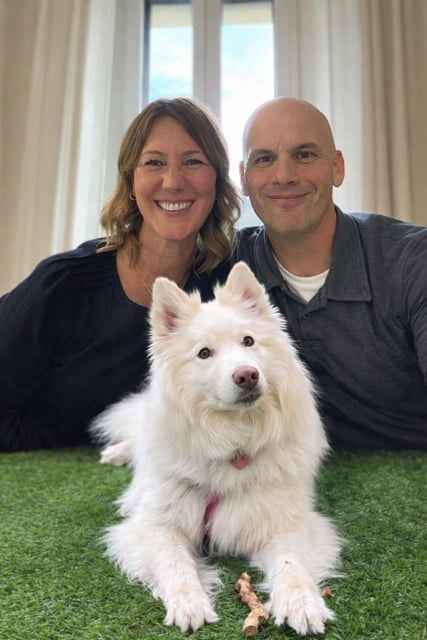 A smiling couple with a white fluffy dog lying on artificial grass in front of a window with sheer curtains