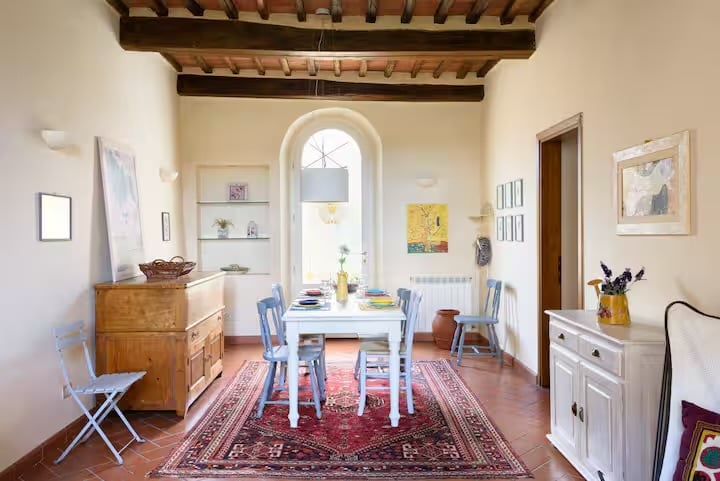 Bright rustic dining room with arched doorway, wooden beams, terracotta floors, red patterned rug, blue chairs, and vintage furnishings