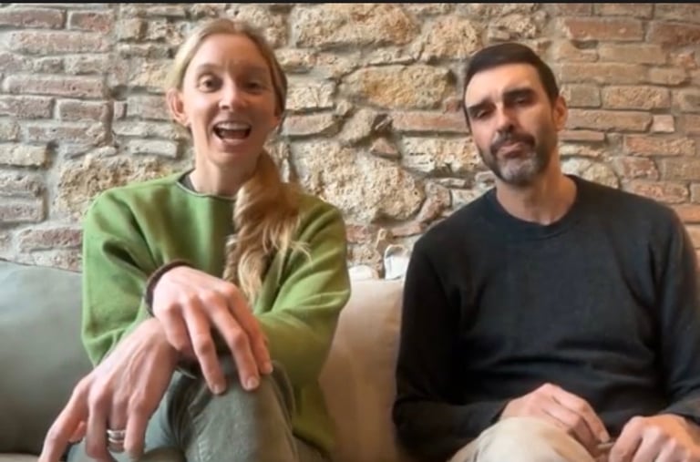 Two people sitting against a brick wall, one wearing a green sweater smiling and one in a black shirt
