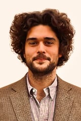 Portrait of a man with dark curly hair and beard wearing a brown plaid blazer and light striped shirt