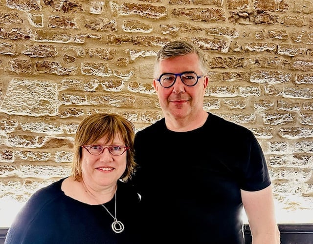 Two smiling people posing together in front of a tan brick wall