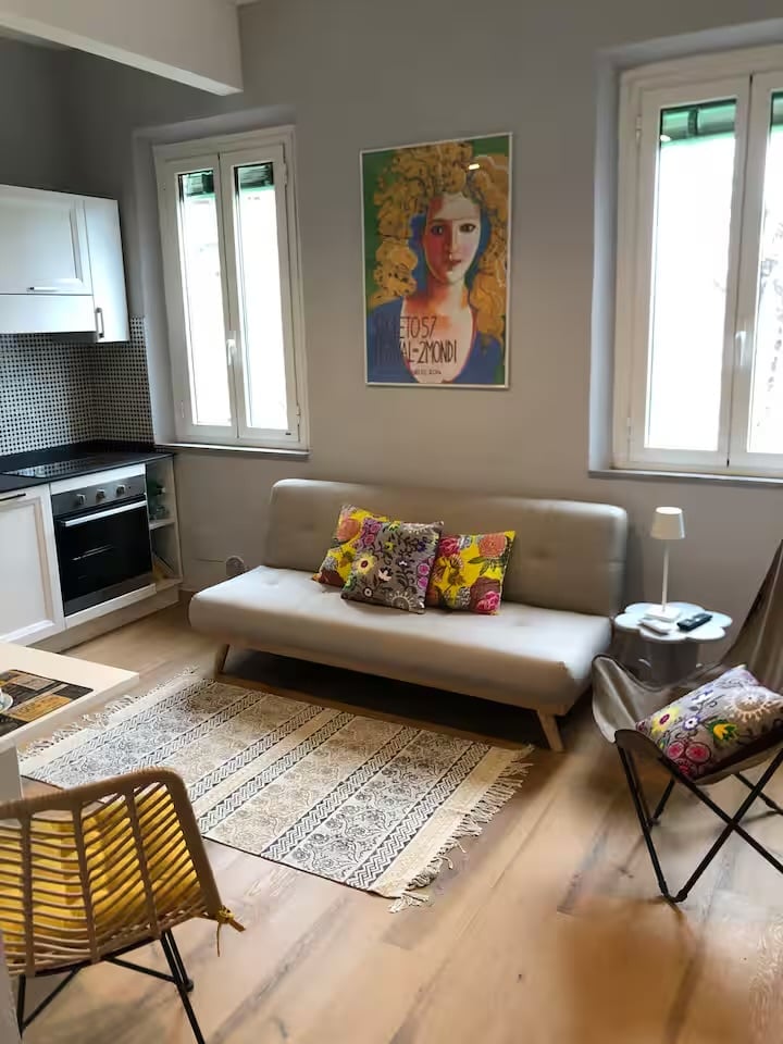 Modern studio apartment with beige sofa, kitchenette, portrait artwork on wall, patterned rug, and wooden flooring