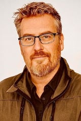 Headshot of a man with reddish-brown hair, glasses, and a beard wearing a brown jacket